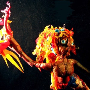 May include: A handmade doll with a red, yellow and blue fabric body. The doll has long, flowing hair made of yellow and red fabric. The doll's eyes are blue and the doll's arms are outstretched.