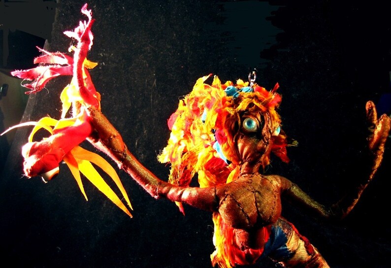 May include: A handmade doll with a red, yellow and blue fabric body. The doll has long, flowing hair made of yellow and red fabric. The doll's eyes are blue and the doll's arms are outstretched.
