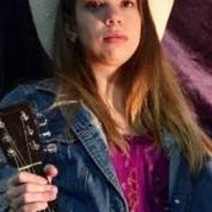 May include: A woman wearing a white cowboy hat and a denim jacket is holding an acoustic guitar. She is wearing a pink and purple shirt underneath the jacket.