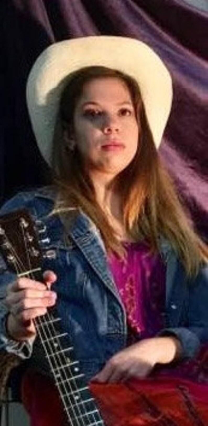 May include: A woman wearing a white cowboy hat and a denim jacket is holding an acoustic guitar. She is wearing a pink and purple shirt underneath the jacket.
