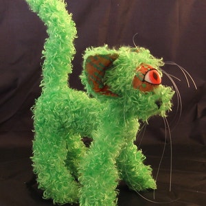 May include: A green, fuzzy, plush toy cat with one red eye and a long, white tail. The cat has a textured, patterned ear.