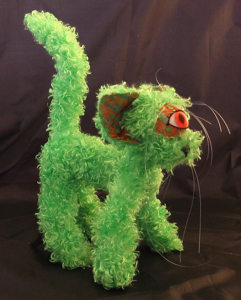 May include: A green, fuzzy, plush toy cat with one red eye and a long, white tail. The cat has a textured, patterned ear.