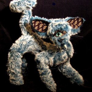 May include: A blue and white furry creature with a long tail and large ears. The ears are decorated with a paisley pattern in gold and black. The creature has a large, toothy grin.