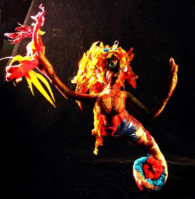 May include: A handmade mermaid doll with a red, yellow and blue fabric body. The doll has long, flowing orange hair and a tail with a red and blue fabric fin. The doll is holding a red and yellow fabric dragon in its hand.