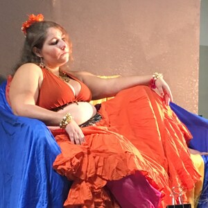 May include: A woman wearing a bright orange skirt and a rust-colored halter top sits on a blue chair. She has a flower in her hair and is wearing gold jewelry.