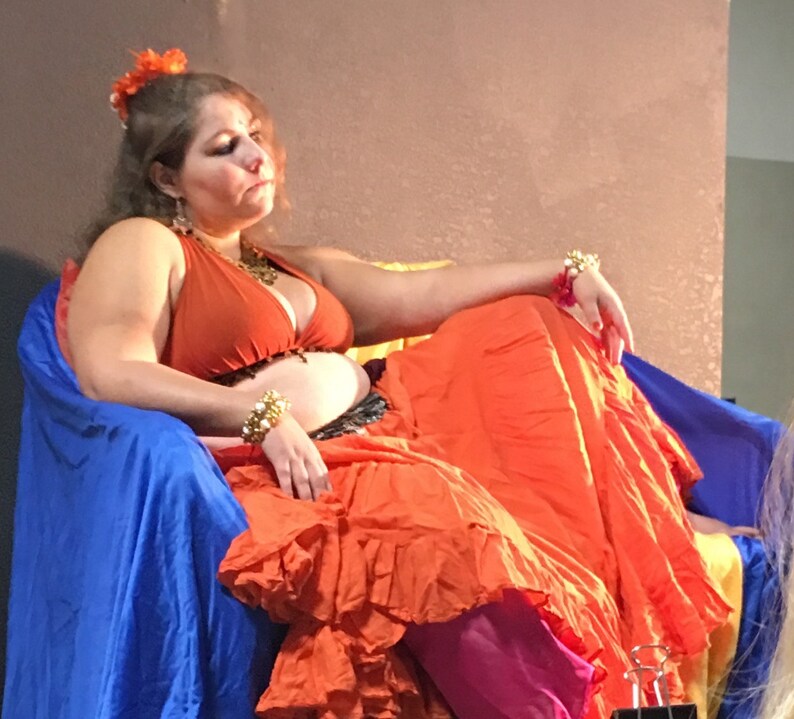 May include: A woman wearing a bright orange skirt and a rust-colored halter top sits on a blue chair. She has a flower in her hair and is wearing gold jewelry.