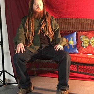 May include: A man with long dreadlocks sits on a wicker couch with red cushions. He is wearing a green jacket, blue jeans, and brown boots. The couch is in front of a red backdrop.