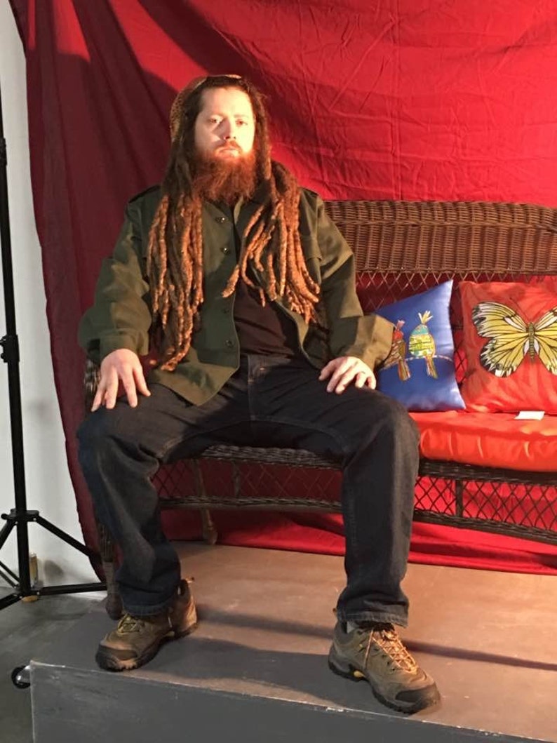 May include: A man with long dreadlocks sits on a wicker couch with red cushions. He is wearing a green jacket, blue jeans, and brown boots. The couch is in front of a red backdrop.