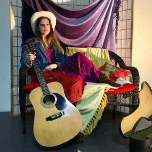 May include: A woman wearing a white cowboy hat and a denim jacket sits on a wicker chair with a guitar in her lap. She is wearing a long, purple and red dress and has a yellow and white blanket draped over her legs. A guitar case is on the floor next to the chair.