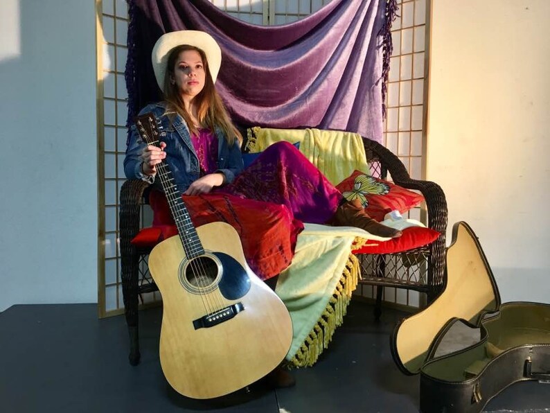May include: A woman wearing a white cowboy hat and a denim jacket sits on a wicker chair with a guitar in her lap. She is wearing a long, purple and red dress and has a yellow and white blanket draped over her legs. A guitar case is on the floor next to the chair.