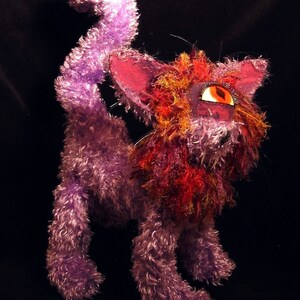 May include: A purple and red furry cat toy with one large, orange eye. The cat has a long, curly tail.