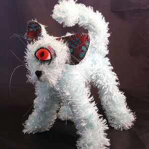 May include: A fluffy, white, plush toy cat with a single large red eye. The cat has blue and green fabric ears with a black nose and mouth.