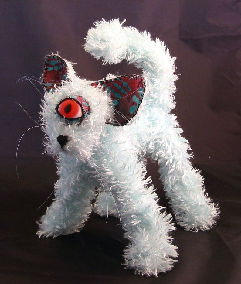 May include: A fluffy, white, plush toy cat with a single large red eye. The cat has blue and green fabric ears with a black nose and mouth.