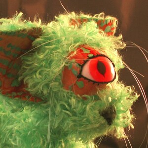 May include: A green, fuzzy, handmade toy with a red eye and a black nose. The toy has a patterned fabric ear and long white whiskers.