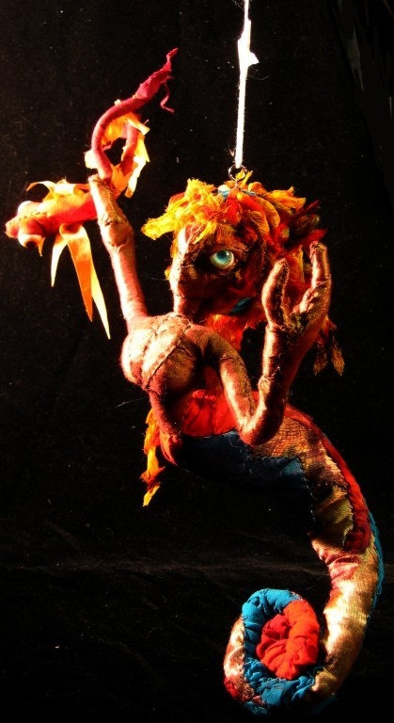 May include: A colourful, handmade fabric doll shaped like a seahorse. The doll is made of red, orange, yellow, blue and white fabric. The doll has a single blue eye and is hanging from a string.
