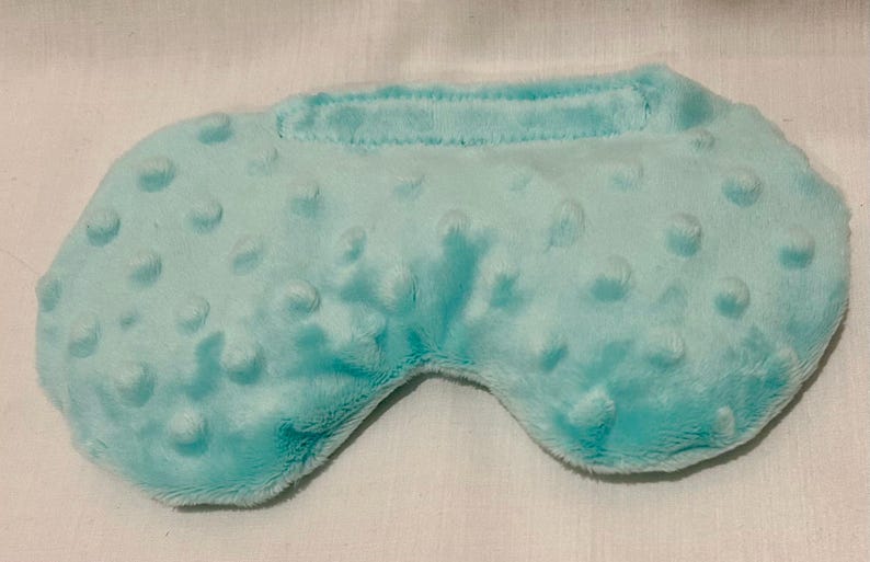 May include: A light blue sleep mask with a textured, dotted surface. The mask has a curved shape to fit around the eyes and a band across the top. The soft material is likely for comfort.