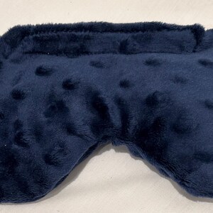 May include: A navy blue sleep mask with a soft, plush texture and a curved shape. The mask has a band across the top and small, raised dots across the surface. The mask is designed to block out light.