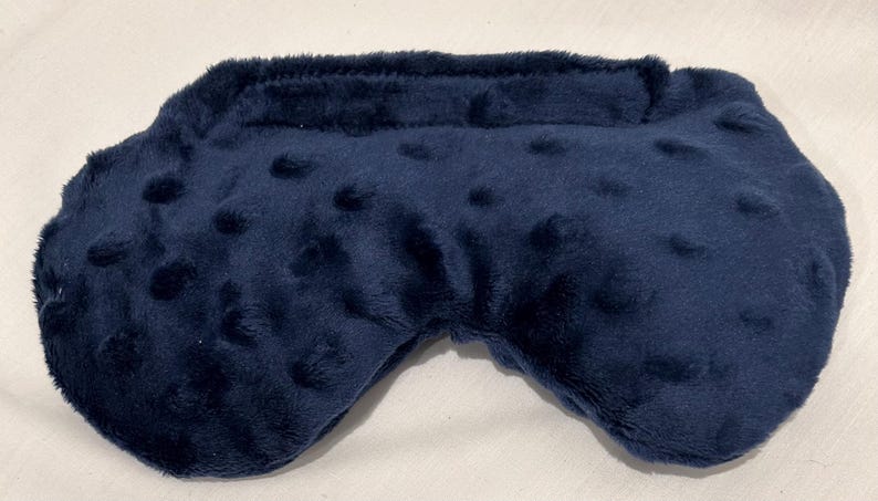 May include: A navy blue sleep mask with a soft, plush texture and a curved shape. The mask has a band across the top and small, raised dots across the surface. The mask is designed to block out light.