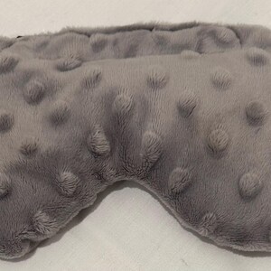 May include: A gray sleep mask with a textured, plush surface. The mask has a rounded shape with a contoured design for the eyes and is made of a soft, fuzzy material. The mask is designed to block out light.