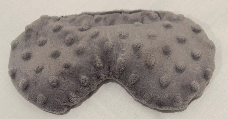 May include: A gray sleep mask with a textured, plush surface. The mask has a rounded shape with a contoured design for the eyes and is made of a soft, fuzzy material. The mask is designed to block out light.