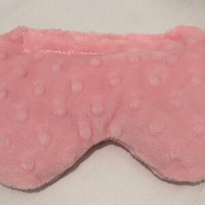 May include: A soft, pink eye mask with a textured, dotted pattern. The mask is designed to cover the eyes and has a curved shape for comfort. The material appears to be a plush fabric.