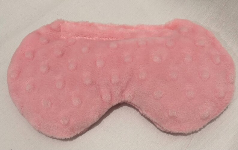 May include: A soft, pink eye mask with a textured, dotted pattern. The mask is designed to cover the eyes and has a curved shape for comfort. The material appears to be a plush fabric.