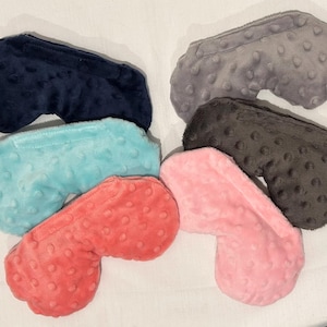 May include: A collection of six plush eye masks in various colors, including navy blue, gray, black, light blue, coral, and pink. Each mask has a textured, dotted surface and a curved shape designed to fit the face.