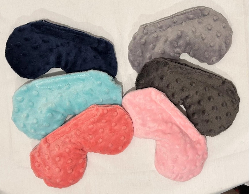 May include: A collection of six plush eye masks in various colors, including navy blue, gray, black, light blue, coral, and pink. Each mask has a textured, dotted surface and a curved shape designed to fit the face.