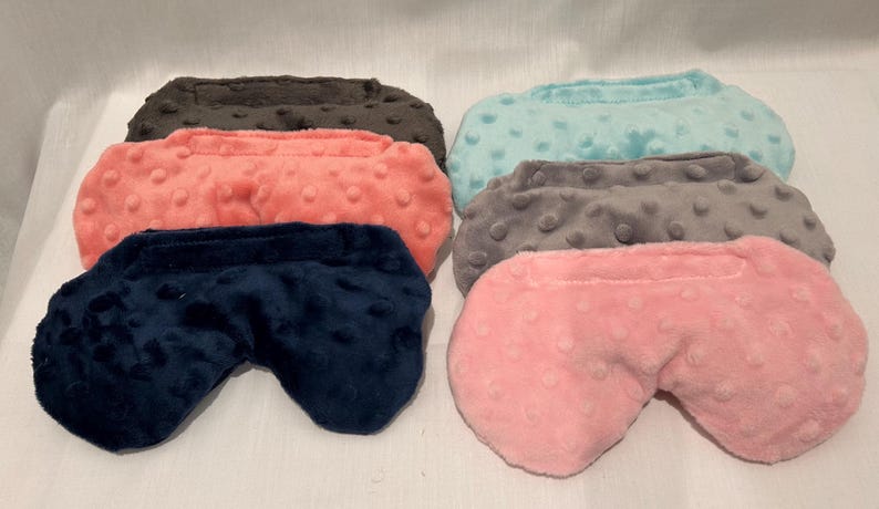 May include: An assortment of six eye masks in various colors. The masks are made of a soft, textured fabric and come in shades of navy blue, coral, pink, gray, teal, and brown. The eye masks are designed to block out light.