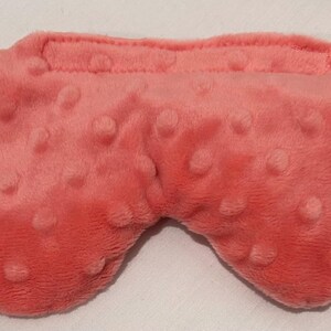 May include: A coral-colored sleep mask with a textured, plush surface. The mask has a rounded shape with a raised section for the nose and a strap at the top. The surface is covered in small, raised bumps.