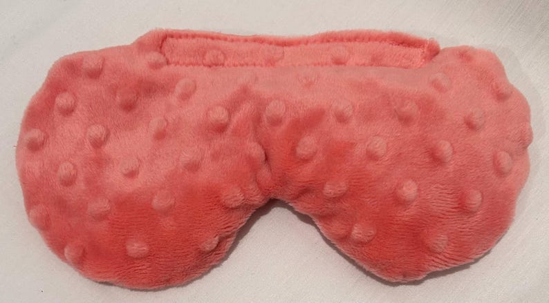 May include: A coral-colored sleep mask with a textured, plush surface. The mask has a rounded shape with a raised section for the nose and a strap at the top. The surface is covered in small, raised bumps.