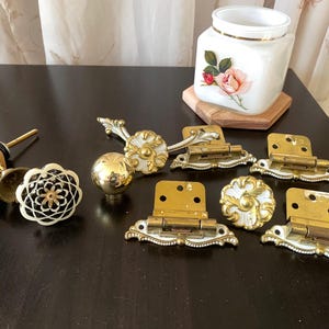 May include: A collection of hardware including two round floral-patterned knobs with white and gold accents, one smooth gold sphere knob, three brass hinges with white decorative edges, and a small bell ornament with a wooden block base.