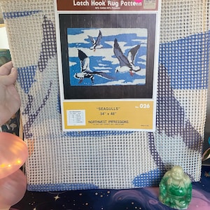 May include: A latch hook rug pattern kit featuring a design of seagulls in flight over a blue and white background. The kit includes the pattern, which measures 34 x 48 inches, and is made of 50% cotton and 50% polyester.