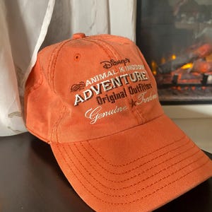 May include: An orange baseball cap featuring embroidered text that reads Disney's ANIMAL KINGDOM ADVENTURE Original Outfitter Genuine Trademark. The cap has a structured crown, a curved brim with visible stitching, and a button on the top center.