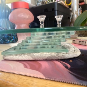 May include: A clear glass tiered stand featuring a flat circular top supported by a square, stepped base composed of multiple stacked glass layers. The structure is transparent with visible green-tinted edges, showcasing a geometric, layered design.