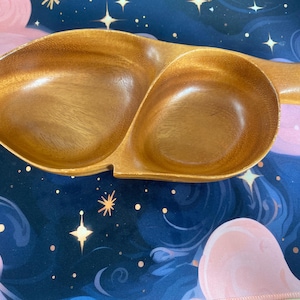 May include: A wooden serving dish with two connected compartments and a handle. The dish is made of light brown wood and has a smooth, polished finish. The design is organic and would be suitable for serving snacks or appetizers.