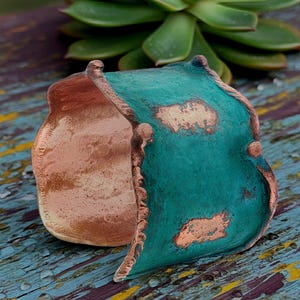 May include: A handcrafted copper cuff bracelet with a distressed turquoise patina. The bracelet has a unique, open design with a textured surface and visible copper accents. The cuff is approximately 2 inches wide.