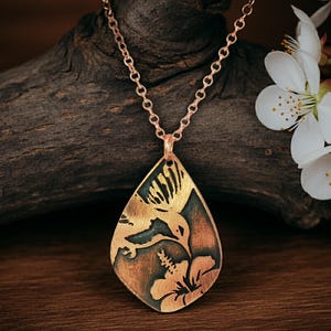 May include: A copper-colored pendant necklace featuring a hummingbird and flower design. The pendant is teardrop-shaped and hangs from a delicate chain. The hummingbird is depicted in flight, with intricate details. The background is a dark, textured wood.