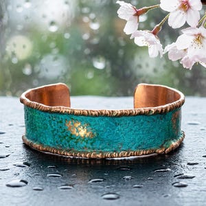 A turquoise-colored cuff bracelet with a copper-colored band sits on a wet surface, with delicate white cherry blossom flowers in the background.