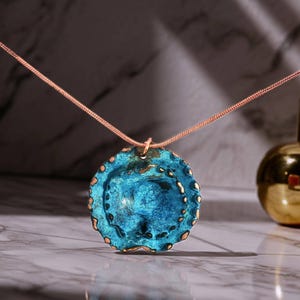 以下が含まれることがあります： 鮮やかなブルーとブロンズ色の縁取りが施された、丸くテクスチャ加工されたペンダントが特徴的な手作りのペンダントネックレス。ペンダントは繊細なローズゴールド色のチェーンから吊り下げられています。ネックレスは大理石の表面に展示されています。