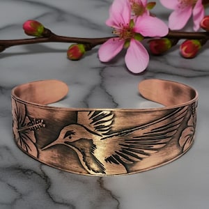 May include: Copper cuff bracelet with a hummingbird and floral design. The open cuff bracelet is a warm copper color with a detailed relief of a hummingbird in flight and flowers. The bracelet is approximately 2.5 inches wide.