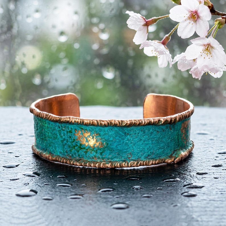 A turquoise-colored cuff bracelet with a copper-colored band sits on a wet surface, with delicate white cherry blossom flowers in the background.