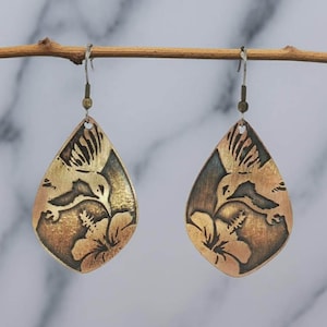 May include: A pair of teardrop-shaped earrings made of antiqued copper. Each earring features a detailed etched design of a hummingbird hovering over a hibiscus flower. The metal has a warm, burnished finish with dark recessed areas to highlight the intricate engraved patterns. The earrings hang from simple metal ear wires.