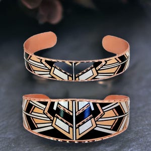 May include: Two copper cuff bracelets featuring a geometric pattern with black, white, and metallic copper enamel inlay. The bracelets have an open back design and a polished finish, showcasing symmetrical angular lines and repeating shapes.