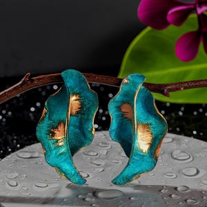 May include: A pair of earrings shaped like curved leaves with a textured teal blue finish and metallic gold accents. The earrings are suspended from a thin brown branch, showcasing a wavy, organic form with a vibrant colour contrast.