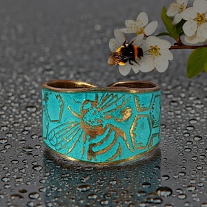 May include: A wide metal ring featuring a textured turquoise patina finish with a raised gold-toned bee and honeycomb pattern. The band has a curved top edge and a metallic base color visible through the worn turquoise surface detailing.
