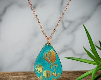 May include: A teardrop-shaped pendant necklace featuring a textured turquoise surface with a gold-colored floral design. The pendant hangs from a delicate rose gold-colored chain, showcasing a botanical motif with three distinct flower buds.