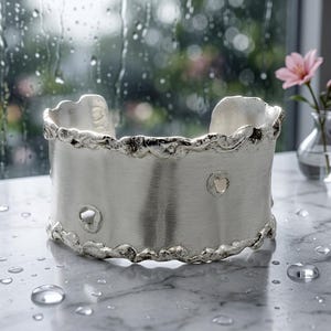 May include: A wide silver cuff bracelet with a brushed finish and an irregular, textured edge. The band features two small, circular cutouts positioned asymmetrically on the front surface, creating a distinct, organic appearance.