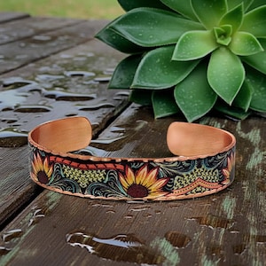 May include: A copper cuff bracelet featuring a repeating pattern of yellow and orange sunflowers set against a dark background with intricate teal and gold filigree details. The metal band has a smooth, polished interior and a textured edge.