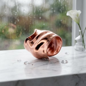May include: A wide cuff bracelet made of polished copper with a distinct, organic, and undulating surface texture. The metal has a warm, metallic sheen that reflects light across its irregular, flowing curves and open-ended circular design.